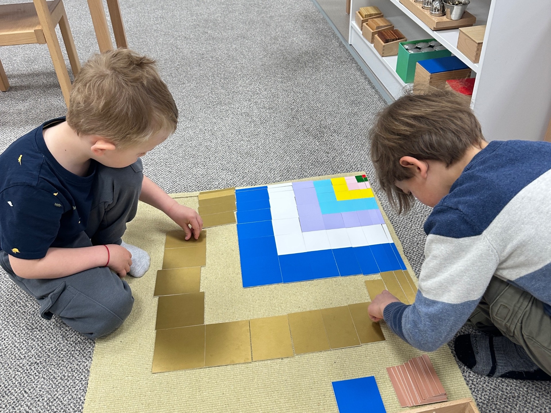 Dva chlapci pracují s montessori matematickou pomůckou barevných čtverců na světlém koberečku.